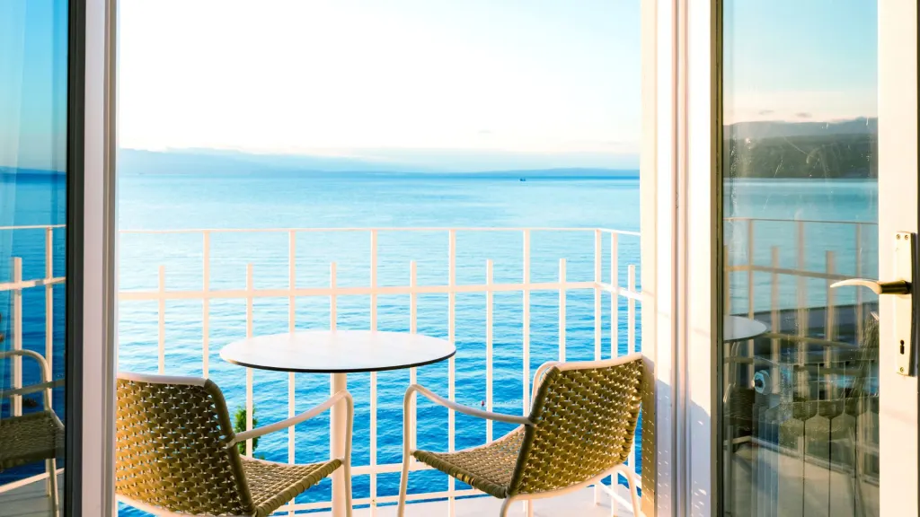 A balcony overlooking the ocean with two wicker chairs and a small round table, framed by open sliding glass doors.